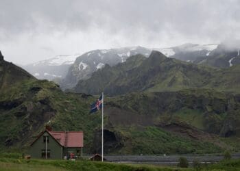 white concrete house with white, blue, and red cross flag near mountain range covered in green vegetation at daytime