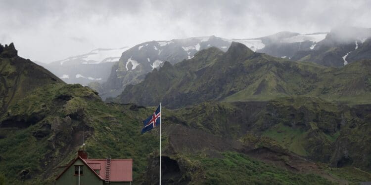 white concrete house with white, blue, and red cross flag near mountain range covered in green vegetation at daytime