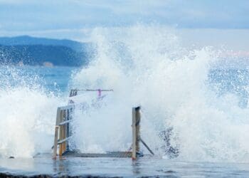 Huge powerful storm waves completely cover the pier, sweeping up splashes of snow-white sea foam. The splashing of huge tropical waves at the pier in close-up. Seascape with huge waves, snow-white foam, splashes and riotous elements for the design of articles, profile header, for web design, applications, creativity and inspiration