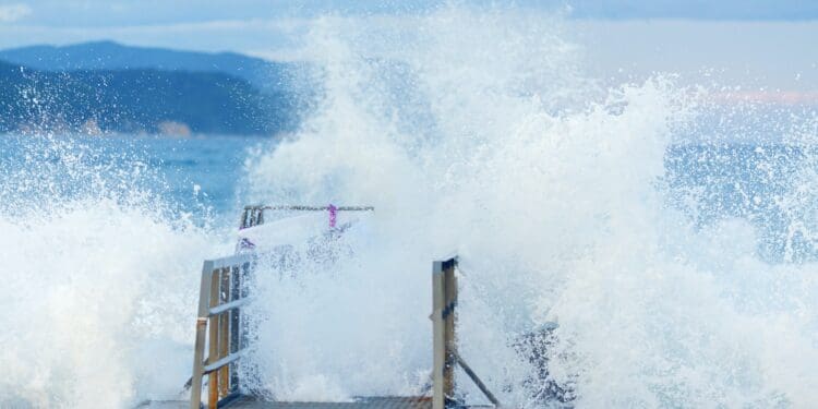 Huge powerful storm waves completely cover the pier, sweeping up splashes of snow-white sea foam. The splashing of huge tropical waves at the pier in close-up. Seascape with huge waves, snow-white foam, splashes and riotous elements for the design of articles, profile header, for web design, applications, creativity and inspiration