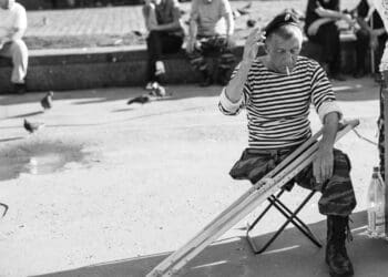 grayscale photo of man sitting on folding stool with crutches in front of people