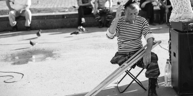 grayscale photo of man sitting on folding stool with crutches in front of people