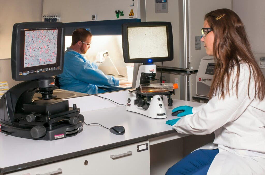A technician viewing a blot on a fluorescence microscope while another technician is using a pipette at the Advanced Technology Research Facility <noread>(ATRF)</noread>, Frederick National Laboratory for Cancer Research, National Cancer Institute.