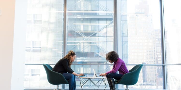 two women sits of padded chairs while using laptop computers