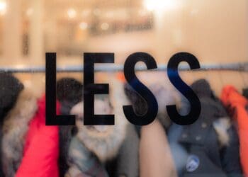 The word “less” on a window, in focus with clothes behind it. Commentary on consumerism and waste in the fashion industry.