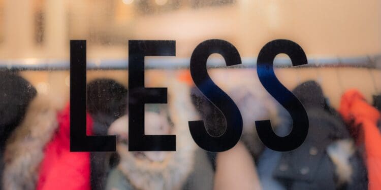 The word “less” on a window, in focus with clothes behind it. Commentary on consumerism and waste in the fashion industry.